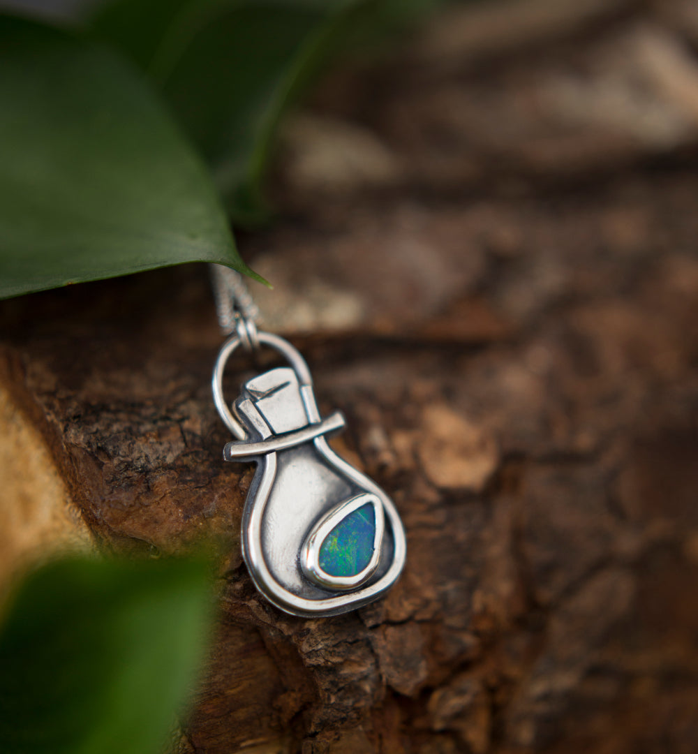 A sterling silver pendant shaped like a small potion bottle with a blue-green iridescent opal doublet, resting on a textured piece of wood with blurred green leaves in the foreground.