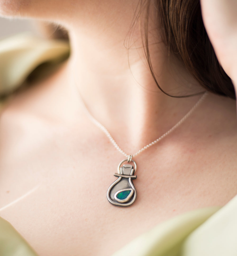 A close-up lifestyle shot of a person wearing a silver pendant necklace with a small green teardrop-shaped opal doublet, set against a soft-focus background.