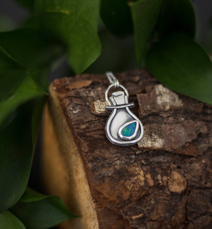 A silver pendant shaped like a potion bottle with a small iridescent blue-green opal doublet, resting on a piece of textured wood surrounded by green leaves.