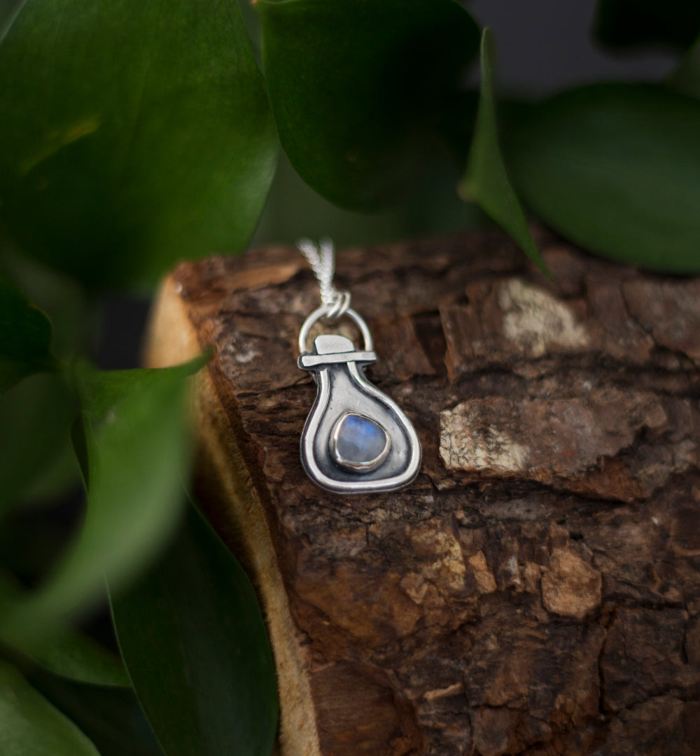 A sterling silver pendant with a potion-bottle shaped setting and a small round rainbow moonstone in the center, resting on a textured piece of wood surrounded by green leaves.