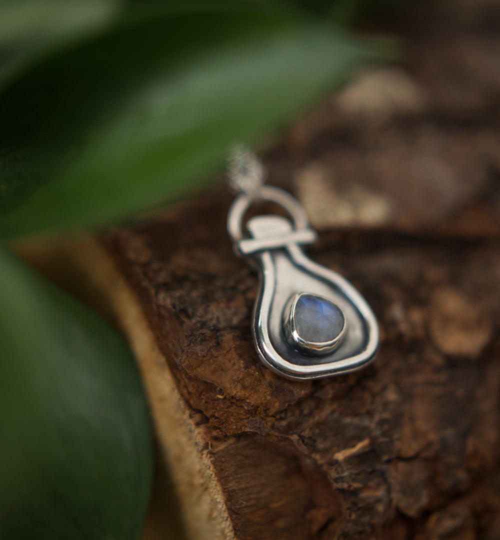 A sterling silver pendant necklace with a potion-bottle shaped setting and a small round moonstone inset, resting on a textured piece of wood with blurred green leaves in the background.