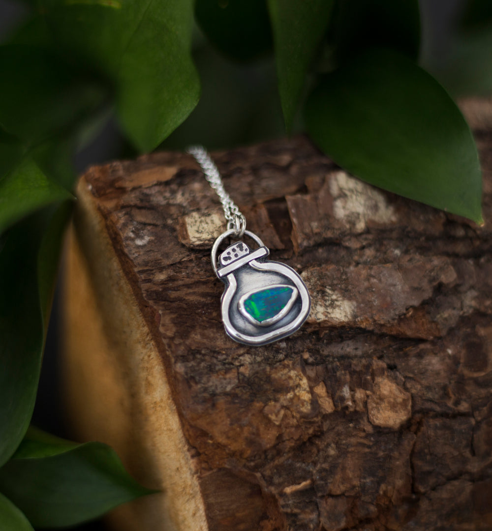 A silver pendant necklace with a small green opal doublet, resting on a piece of textured wood with green leaves in the background.