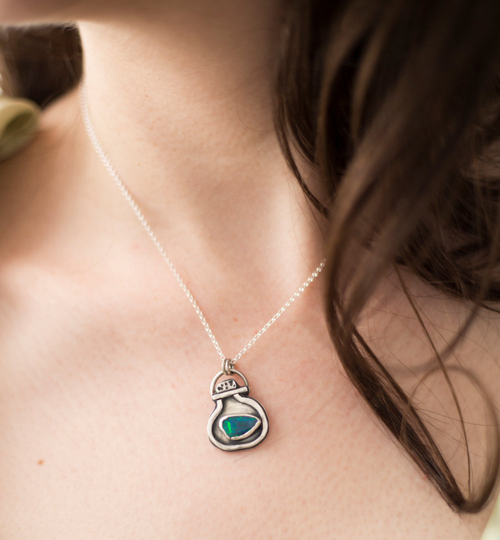 A close-up of a person's neck and chest wearing a delicate silver chain necklace with a small pendant featuring a green and blue opal doublet in a silver pendant.