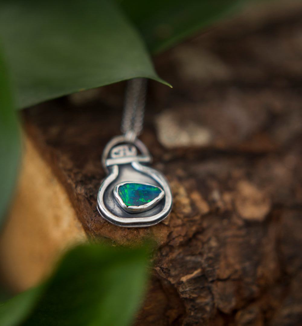 A sterling silver pendant necklace featuring a teardrop-shaped green opal doublet gemstone, resting on a textured piece of wood with blurred green leaves in the foreground.