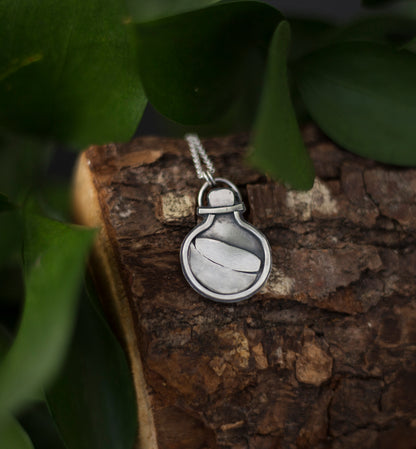 A silver pendant necklace shaped like a small potion bottle, resting on a piece of textured wood with green leaves in the background.