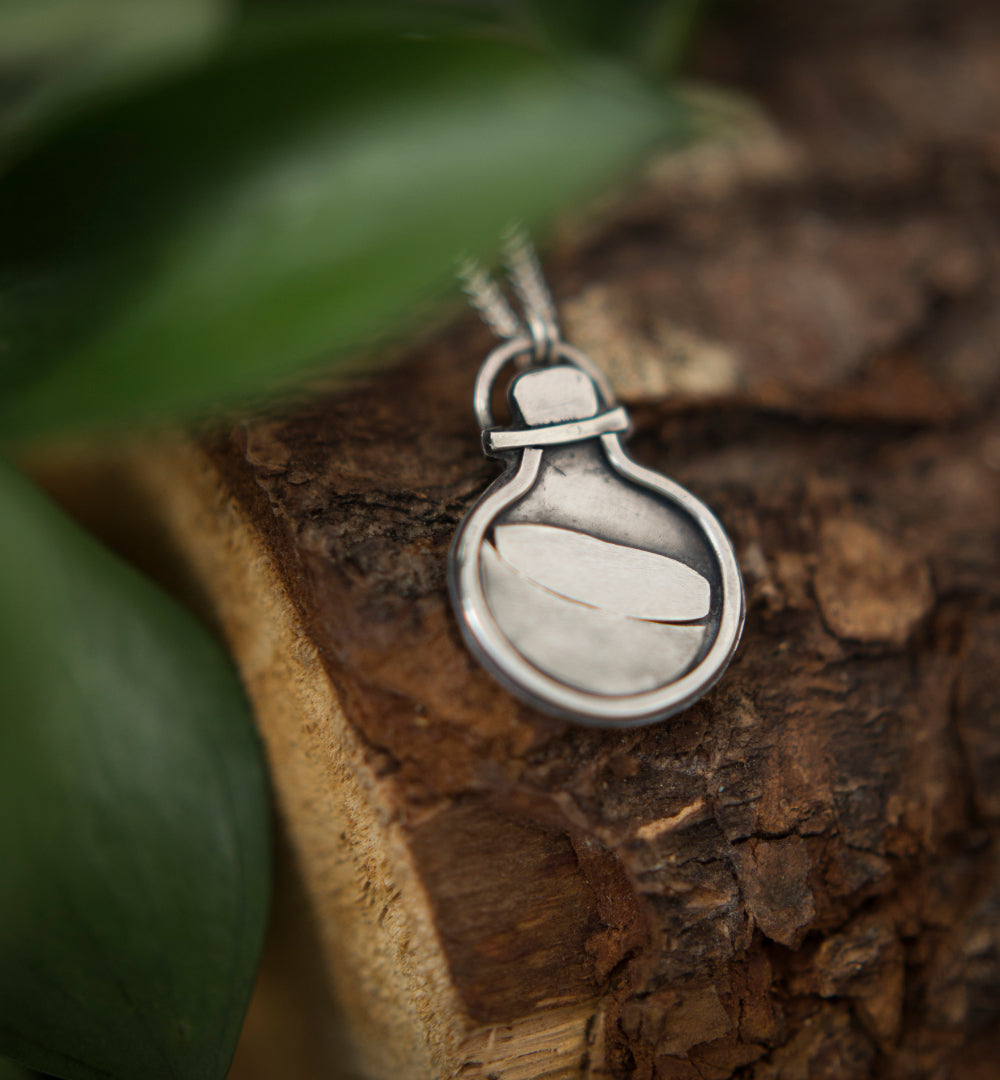A silver pendant necklace featuring a stylized potion bottle design, resting on a piece of textured wood with blurred green leaves in the background.
