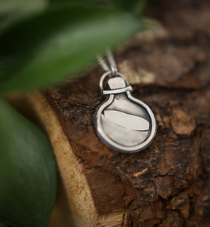 A silver pendant necklace featuring a stylized potion bottle design, resting on a piece of textured wood with blurred green leaves in the background.