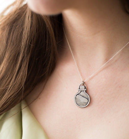 A close-up of a person wearing a delicate silver chain necklace with a small potion bottle shaped pendant