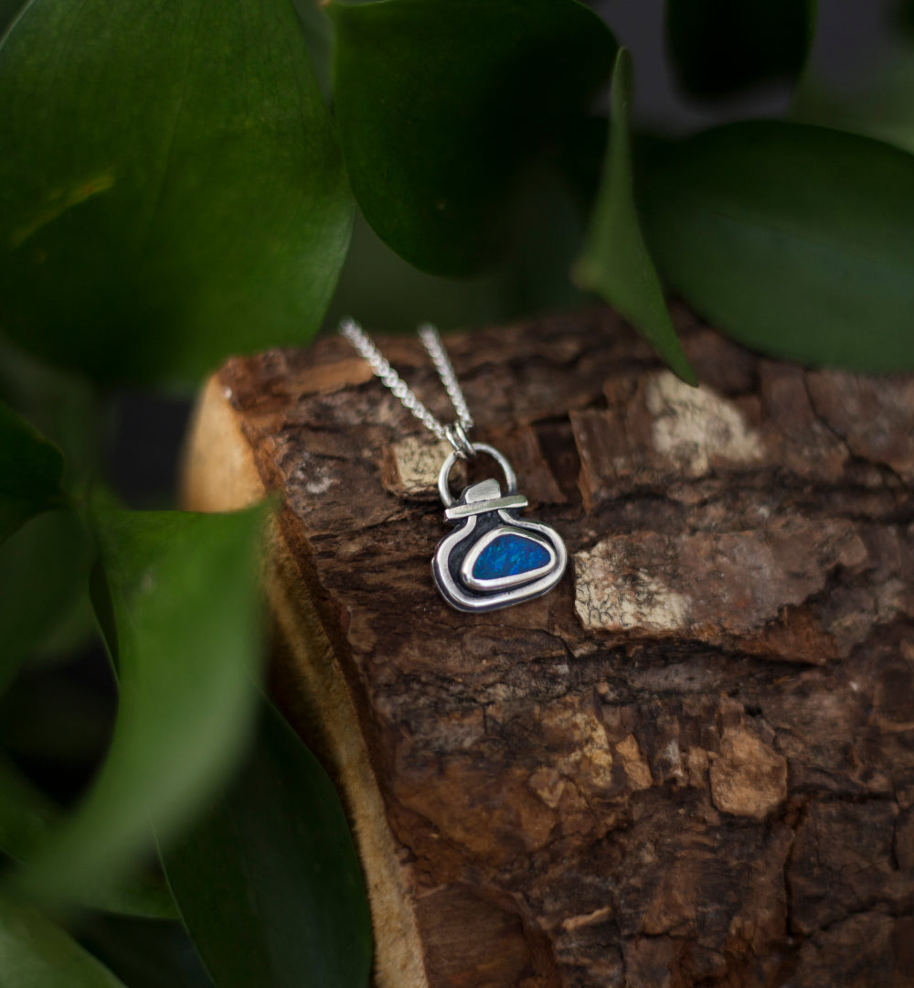 A silver pendant necklace with a blue opal doublet, resting on a piece of textured tree bark surrounded by green leaves.