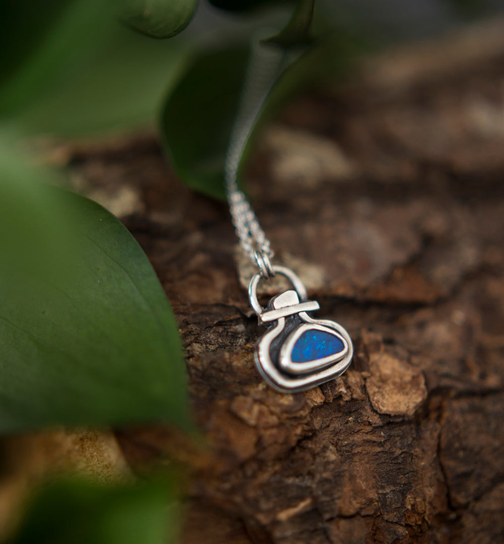 A silver pendant necklace with a small  blue opal doublet, resting on textured brown bark with green leaves blurred in the foreground.
