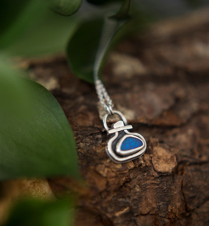 A silver pendant necklace with a small  blue opal doublet, resting on textured brown bark with green leaves blurred in the foreground.