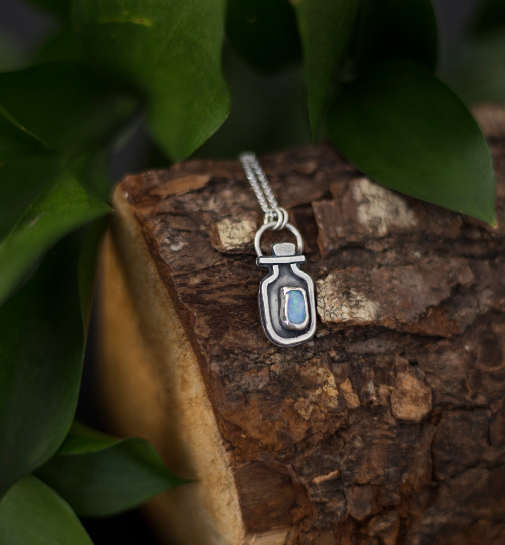 A silver pendant necklace shaped like a small vial with a blue opal doublet, resting on a piece of rough tree bark surrounded by green leaves.