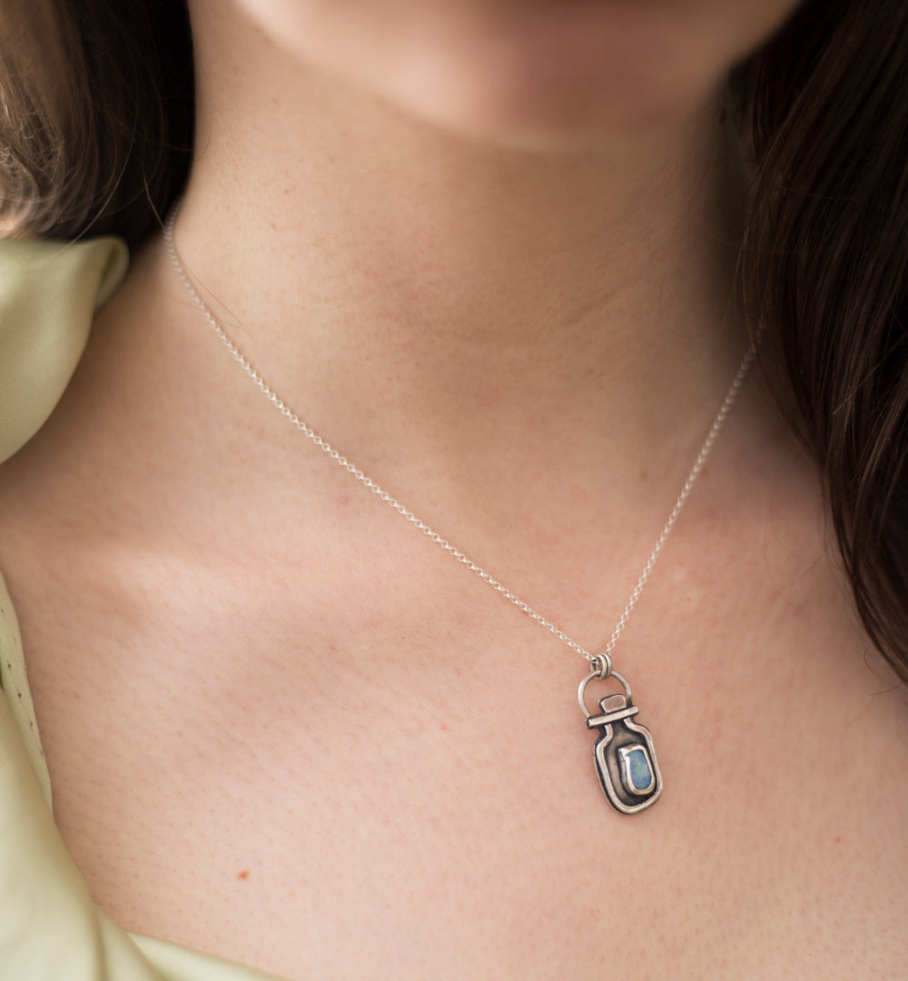 A close-up of a person's neck and chest wearing a delicate silver chain necklace with a small pendant shaped like a vial containing a light blue opal doublet.