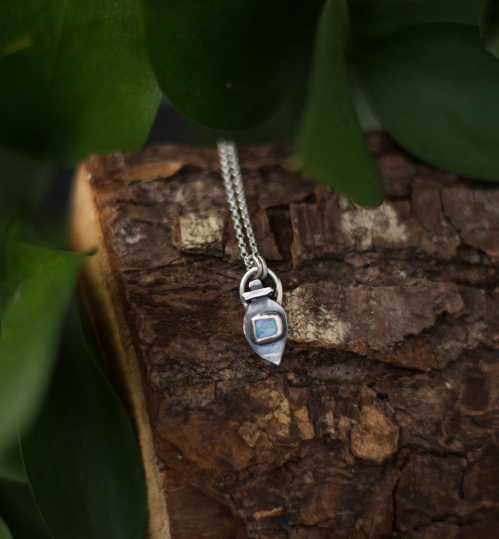 A silver pendant necklace with a small potion-like charm and a light blue opal doublet, resting on a textured piece of wood with green leaves in the background.