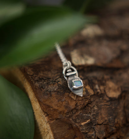 A silver pendant necklace with a small rectangular iridescent blue-green opal doublet, resting on a textured piece of wood with blurred green leaves in the background.