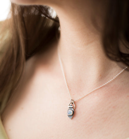 A close-up lifestyle shot of a delicate silver chain necklace with a small pendant worn by a person. The pendant features a silver metal body with a small, light blue square opal doublet.