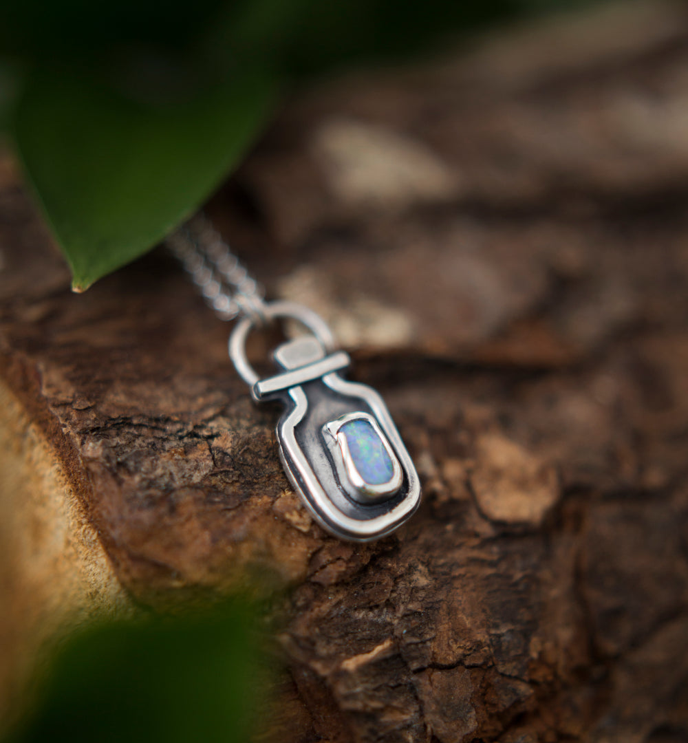 A silver pendant necklace with a small iridescent opal doublet, resting on textured brown bark with blurred green leaves in the background.